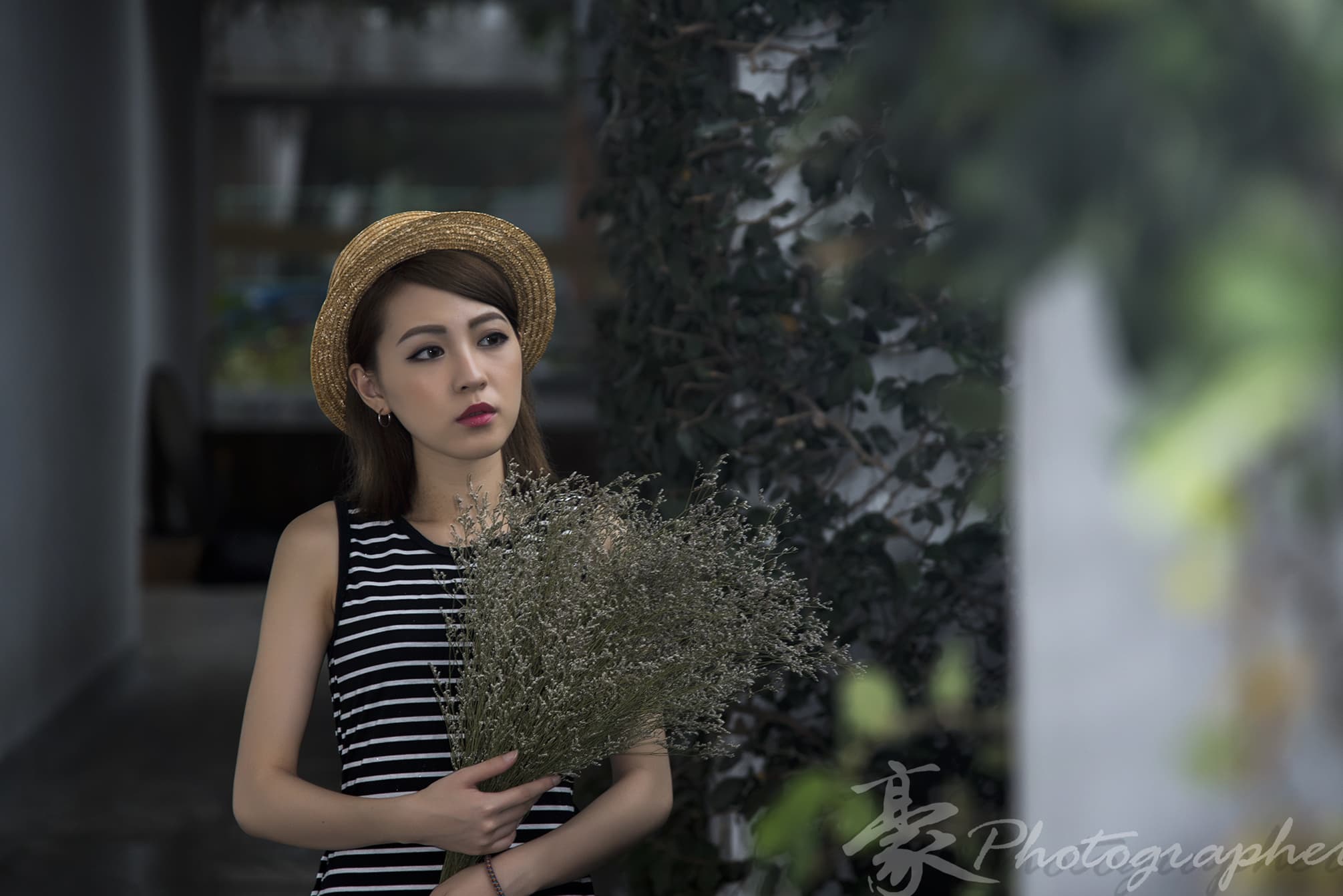 這張照片是一個戴著草帽的女孩站在綠色植物前的照片。她穿著條紋上衣,神情憂鬱,手中拿著一束白色的小花。