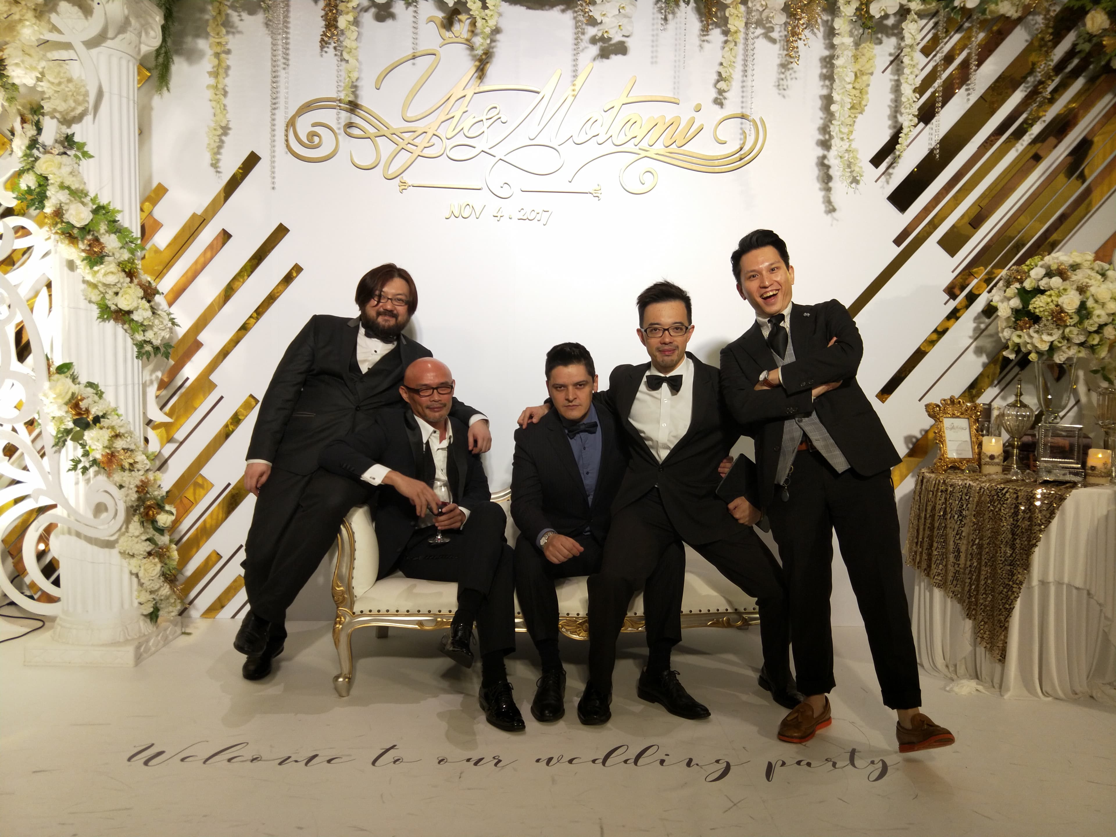 這張婚禮的照片是五位新郎的友人,他們穿著西裝,背景是金色的裝飾,以及花朵裝飾。