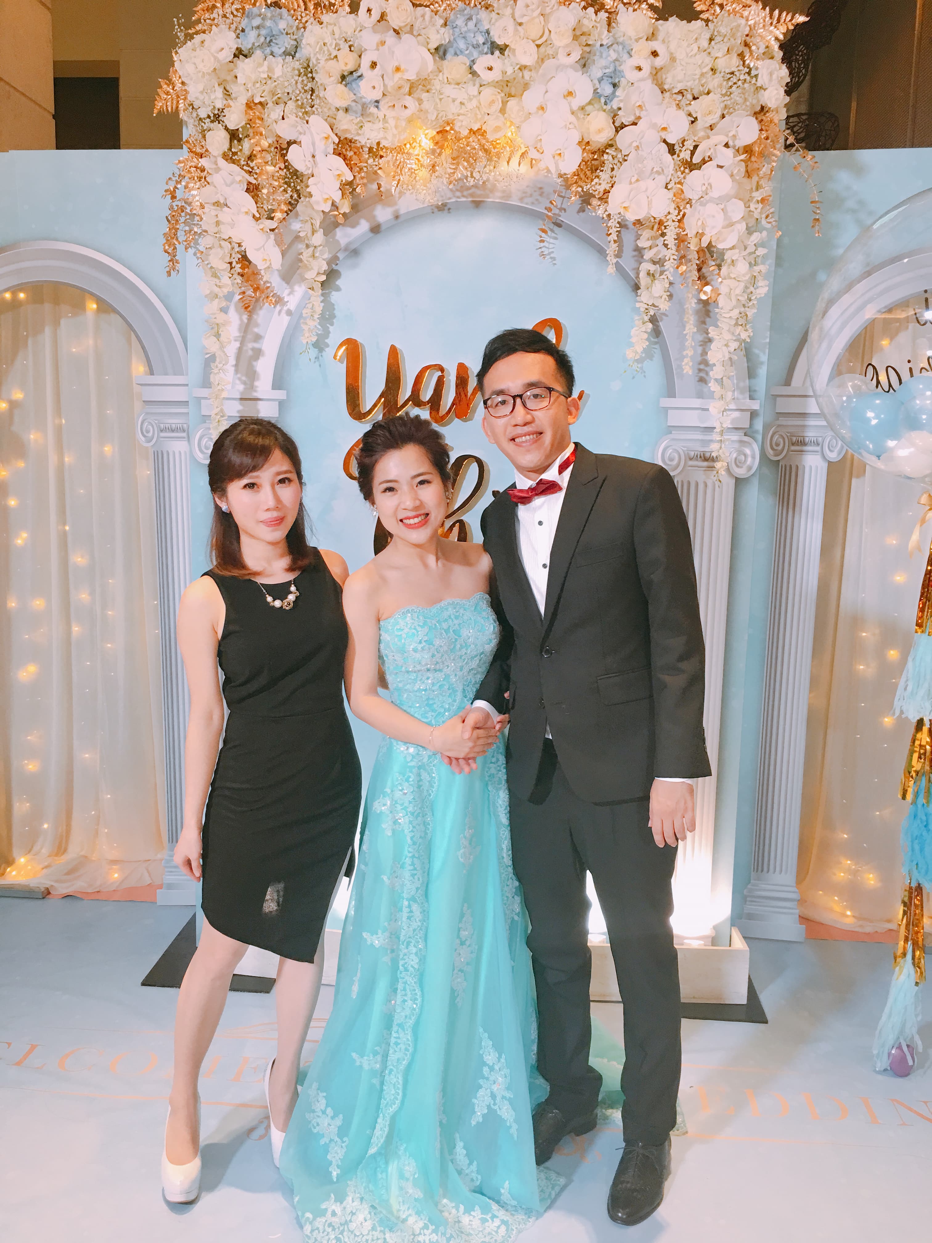 這張「婚禮」的照片是三位新人在一個佈置華麗的婚禮現場合照, 新郎穿著黑色的西裝, 新娘穿著水藍色的禮服, 另一位女性友人則穿著黑色的禮服。