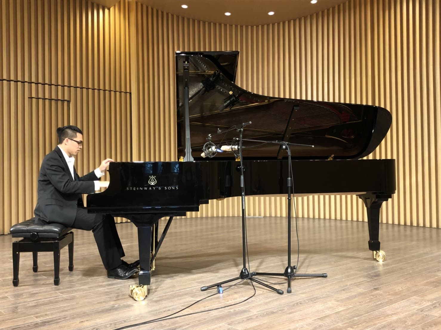 這張「鋼琴演奏」的照片是:一位男士在演奏黑色鋼琴,背景是木質牆面,地板上擺放了兩支麥克風架。