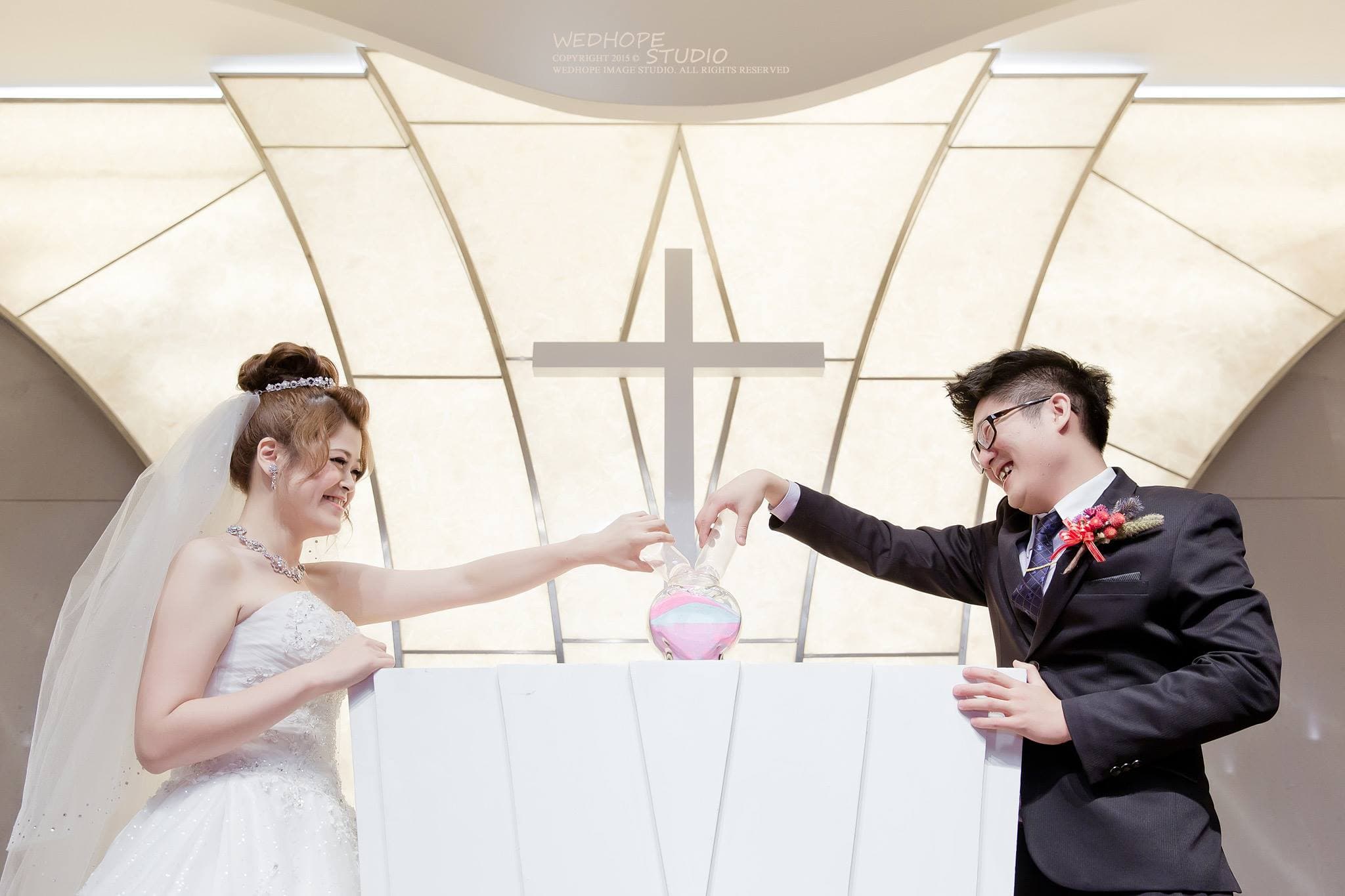 這張照片是一對新人正在教堂裡進行沙畫儀式。新人們手持沙瓶,正準備將沙子倒入一個白色禮盒中,象徵他們將要一起度過美好的未來。他們身後的教堂窗戶透著陽光,照亮了整個儀式現場。