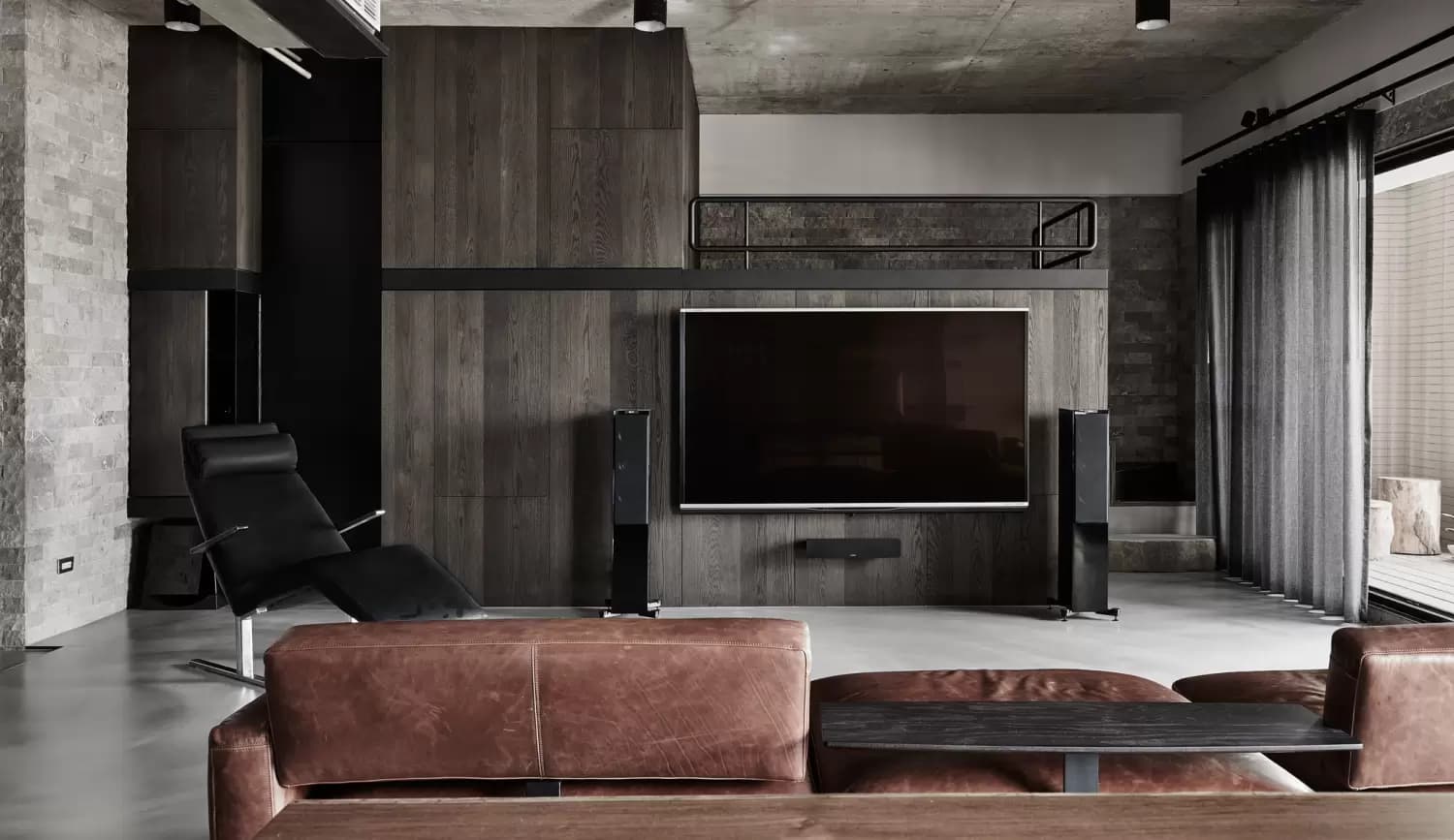這張「客廳」的照片是一個現代化的居家空間,以深色木質和灰色石材為主,配有黑色的休閒椅和棕色皮革沙發。牆上掛著一臺大型電視,下方放置著音響,整體風格沉穩大氣。