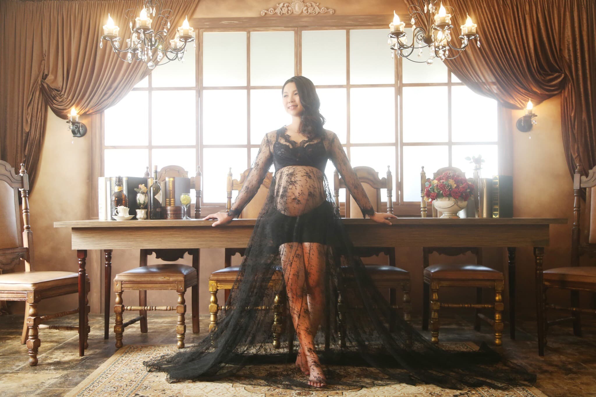 這張照片是一個懷孕的女性站在桌子旁,她穿著一件黑色的蕾絲禮服,禮服的裙擺很長,覆蓋了地板。她看起來很優雅,也很開心。