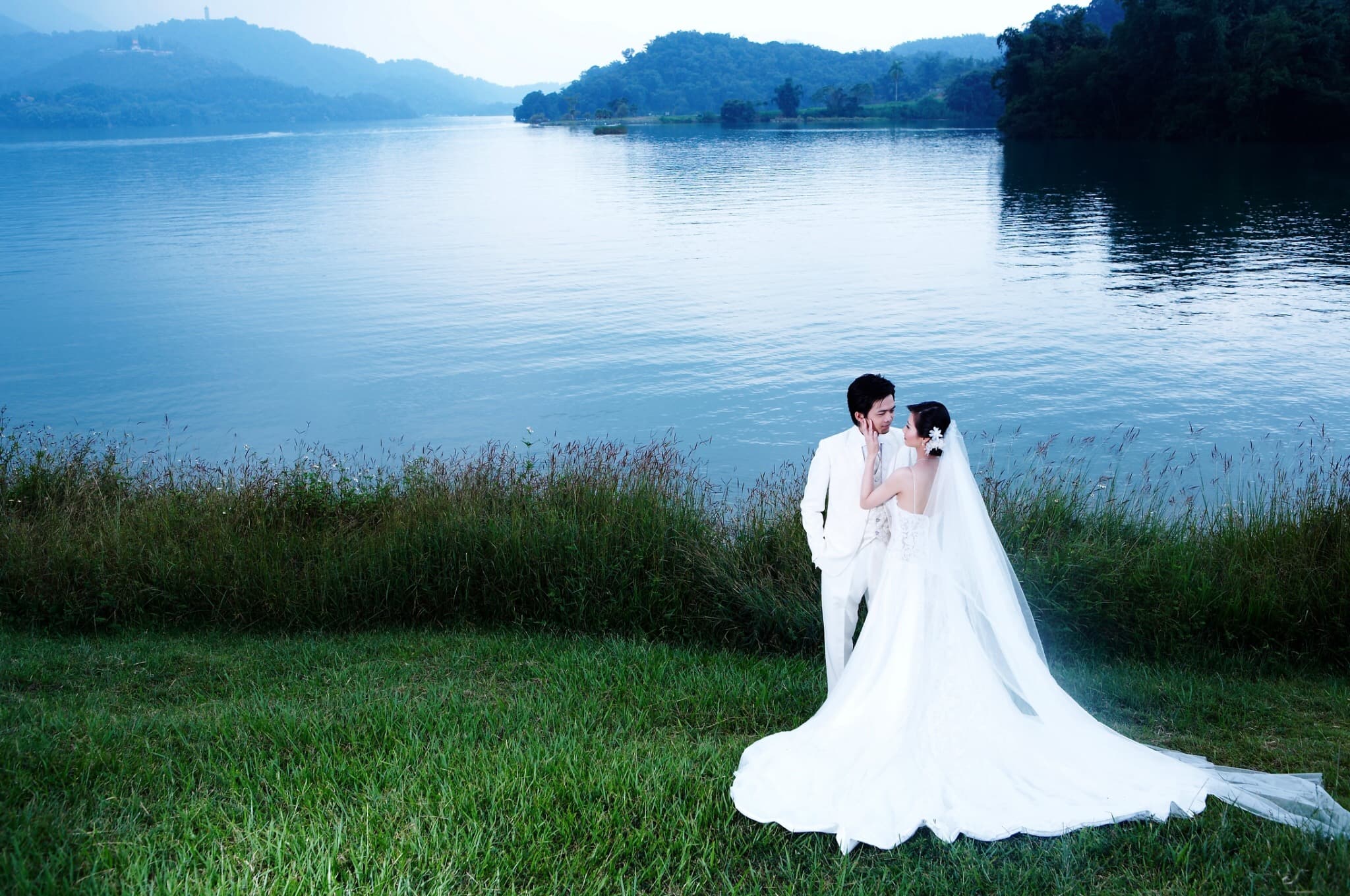 這張「婚紗照」的照片是一對新人站在湖邊,新郎穿著西裝,新娘穿著婚紗,他們互相凝視,背景是湖水和綠色的草地。