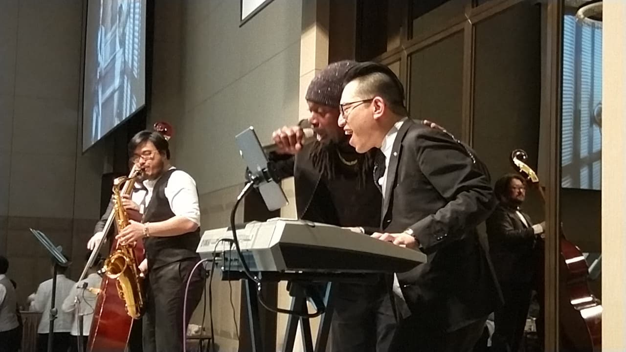 這張音樂表演的照片是三位音樂家在演奏。其中一位演奏薩克斯風,一位彈鍵盤,另一位站在鍵盤旁,似乎正在唱歌。他們身後有一把大提琴,他們看起來正在享受音樂。