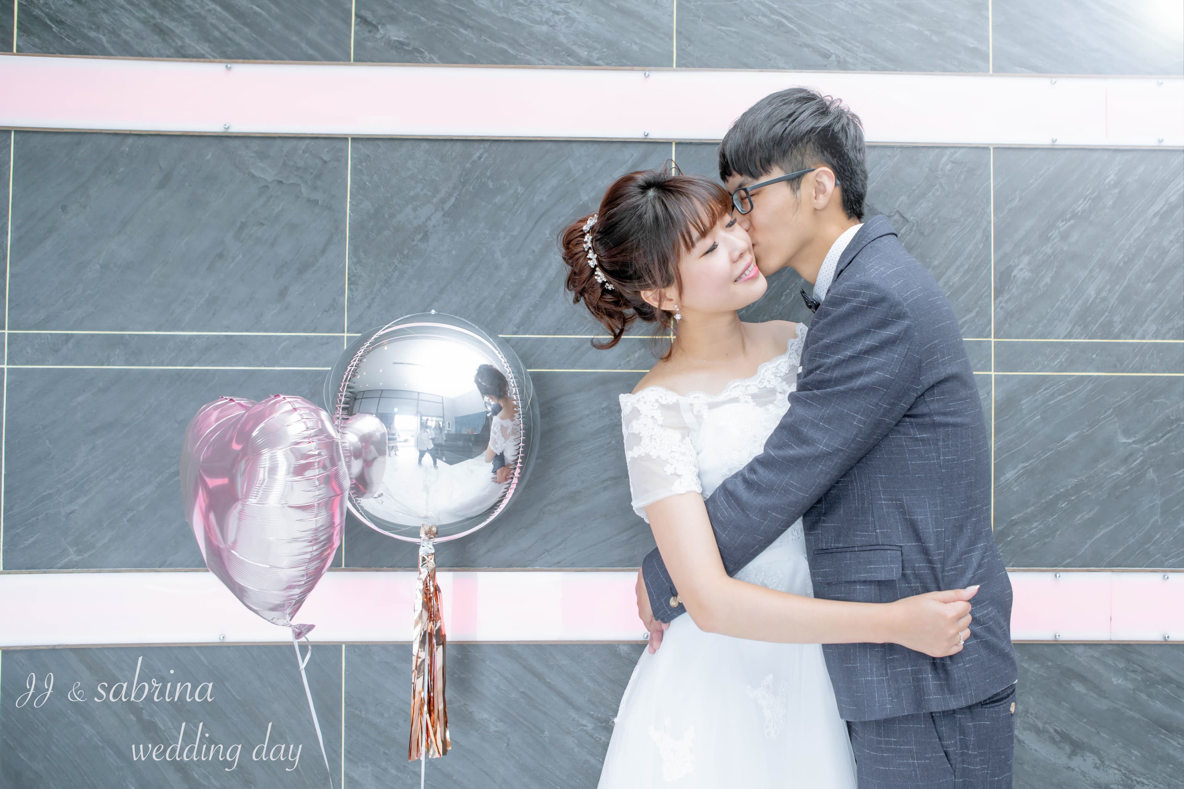 這張「婚禮」的照片是一對新人擁抱在一起,新郎親吻新娘的臉頰,背景是灰色磁磚牆和氣球。