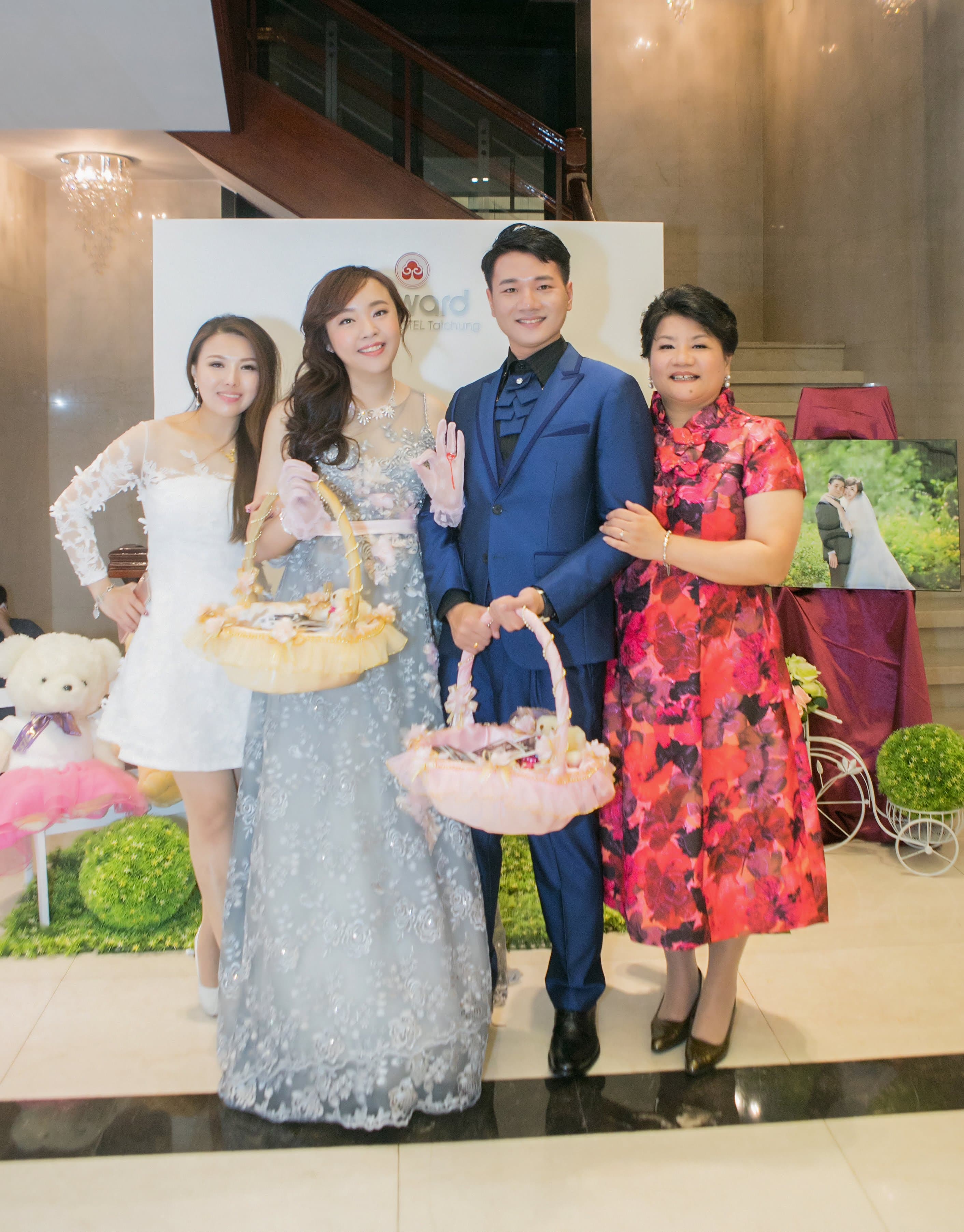 這張照片是三位女性和一位男性站在一起,其中一位女性穿著白色蕾絲連衣裙,另一位女性穿著藍色亮片禮服,第三位女性穿著紅色花卉圖案禮服,男性穿著藍色西裝。他們都帶著笑容,看起來很開心。