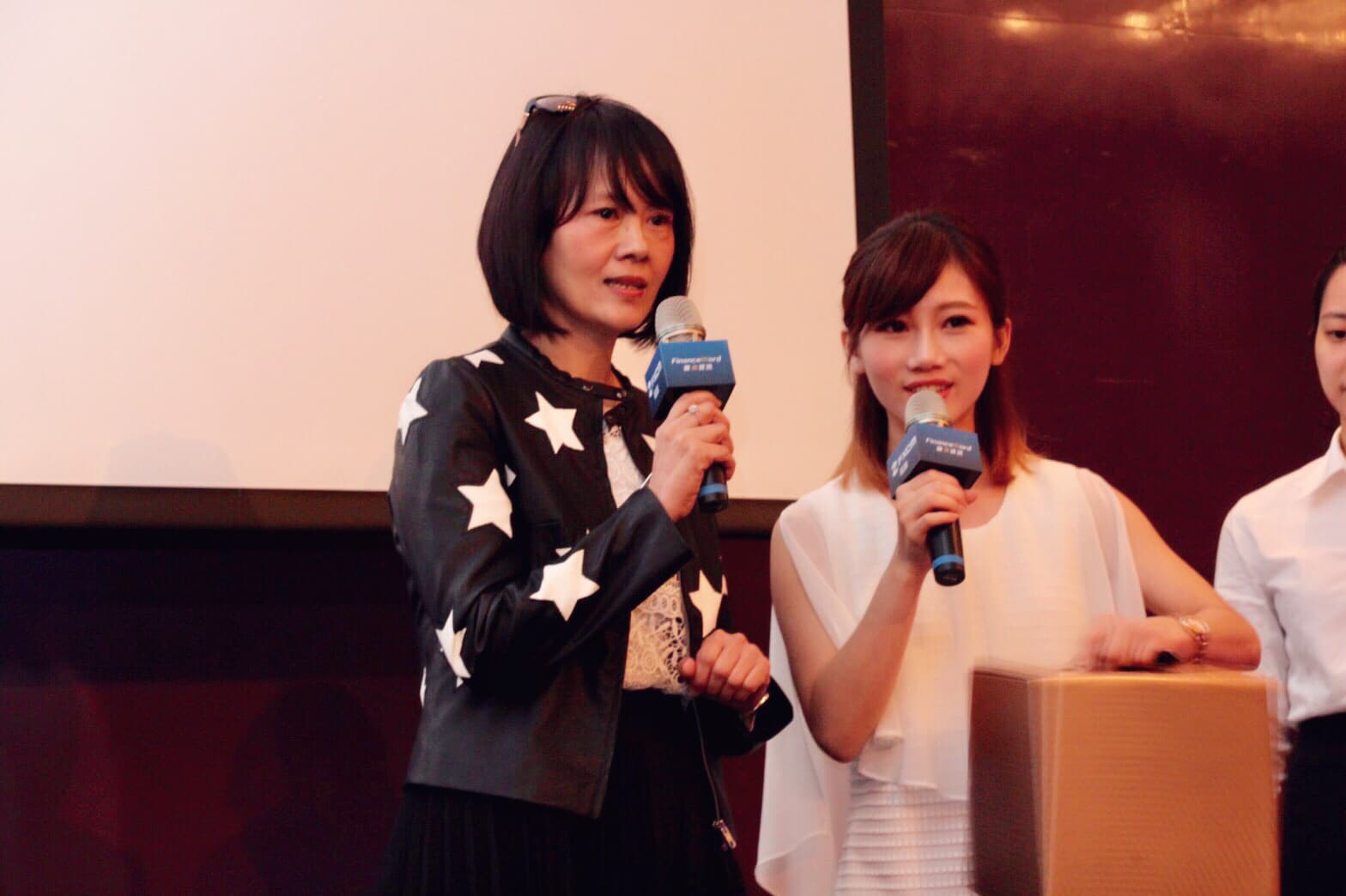 這張照片是三個女人,其中一個女人穿著黑色皮衣,站在最前面,她正在拿著麥克風講話。