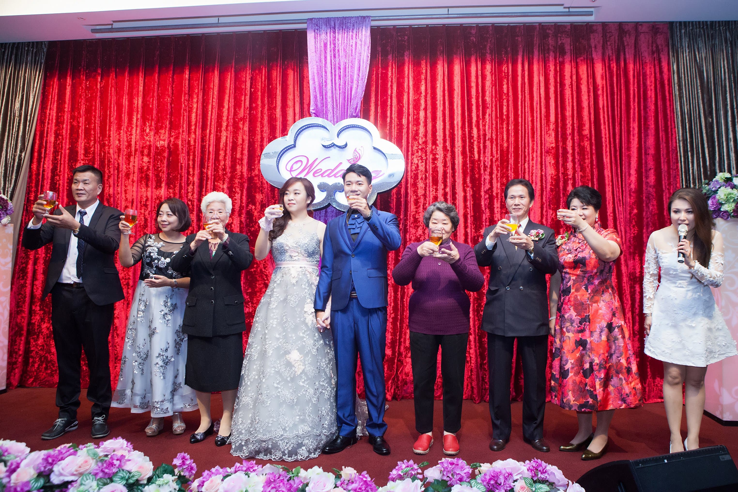 這張照片是一對新人和他們的親朋好友在婚禮上的合照。新人站在中間,穿著婚紗禮服,笑容滿面,非常幸福。親朋好友們圍繞在他們周圍,舉杯祝賀,氣氛熱鬧而溫馨。