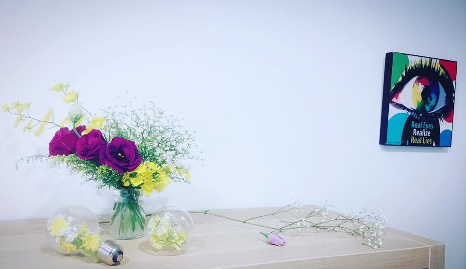 1) 照片顯示一束紫紅色和黃色的花,插在透明花瓶中,放在木製桌子上,旁邊有兩個玻璃燈泡,照片的右上角有一幅畫作,畫作中有一隻眼睛,背景是白色牆壁