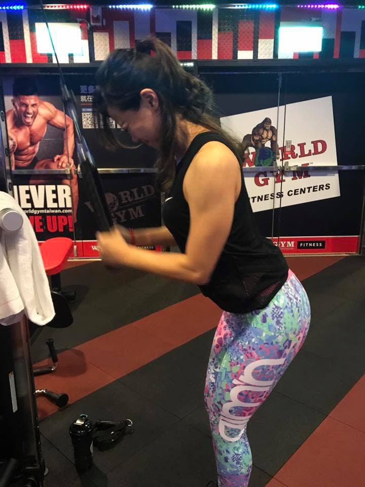 這張照片是一位女士在健身房運動,她穿著鮮豔的彩色緊身褲,正在使用器材鍛煉手臂肌肉。