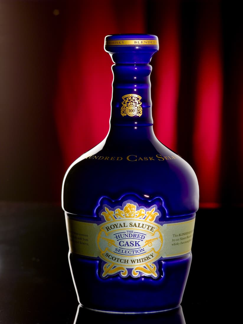 這張「皇家禮炮百年橡木桶威士忌」的照片是, 藍色的酒瓶,瓶身上印著金色文字,瓶身上還有金色圖案,酒瓶背景是一塊深紅色的布。