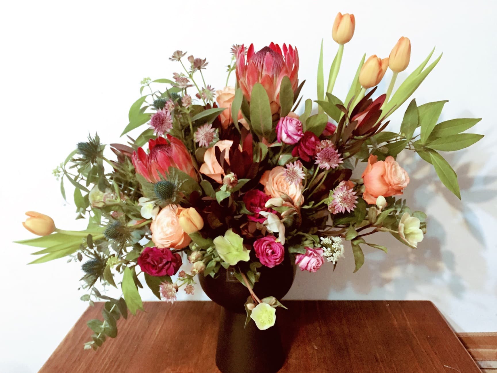 這張花束的照片是一個由不同種類的花卉組成的,包括紅色、橙色和粉色花朵。 其中有紅色的非洲菊,橙色的鬱金香,以及粉色的玫瑰。 它們被插在黑色的花瓶中,擺放在木桌上面。