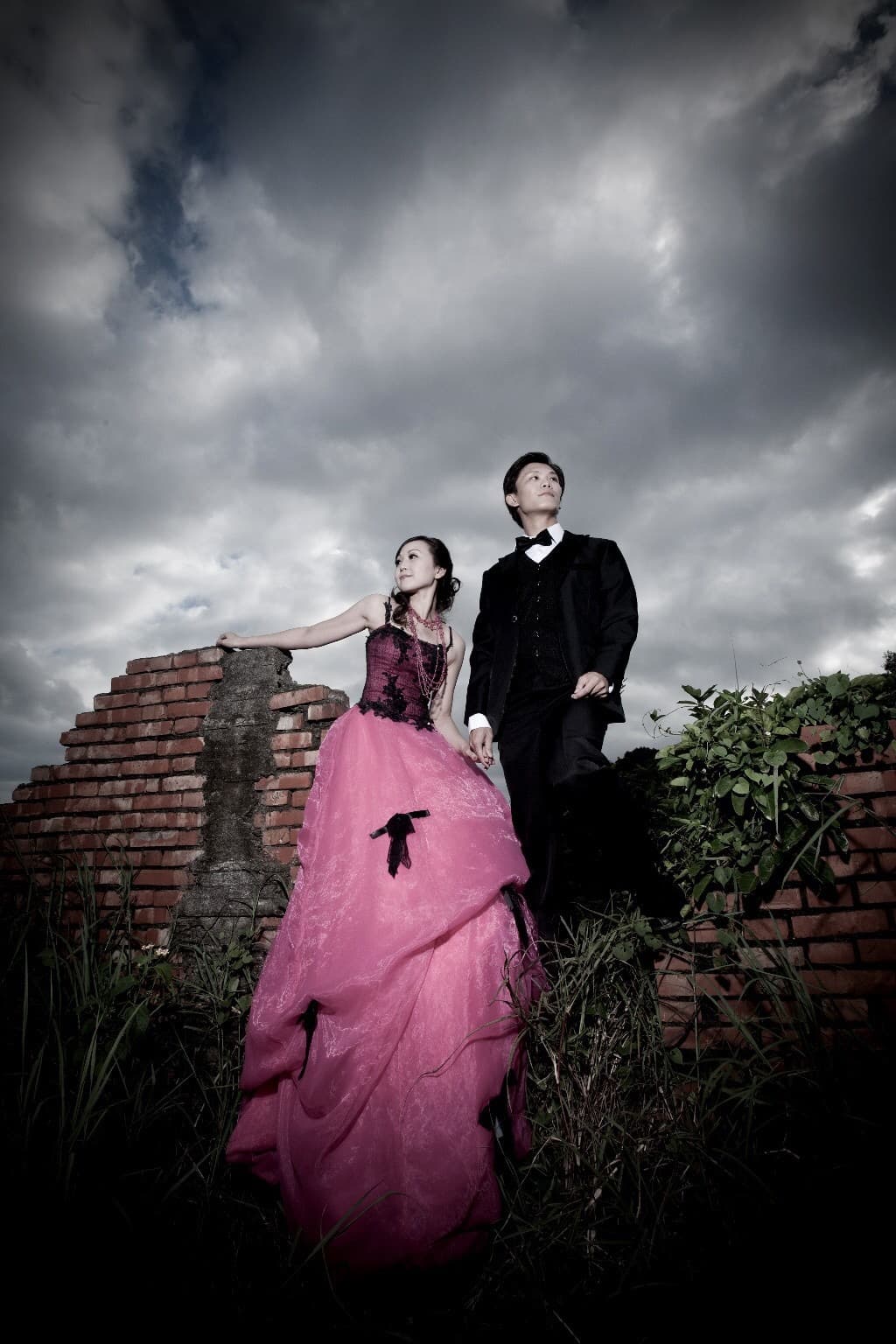 這張婚紗照是一對新人站在紅磚牆前,新娘穿著粉紅色的婚紗,新郎穿著黑色西裝。