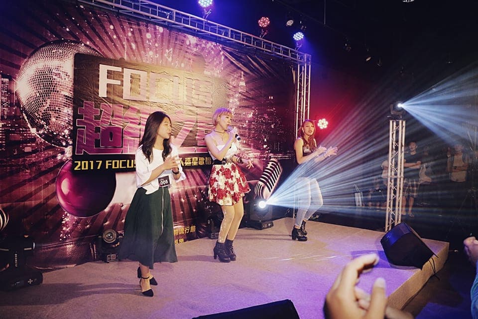 這張照片是三位女歌手在舞臺上演唱。她們穿著亮麗的衣服,手持麥克風,表情充滿活力,正盡情享受演唱的樂趣。背景是五彩繽紛的舞臺燈光,營造出熱鬧的氣氛。