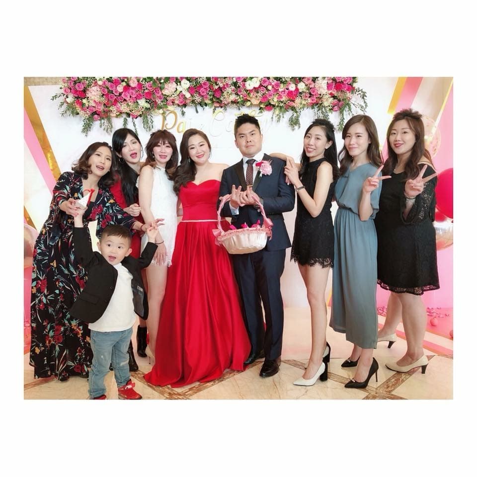 這張照片是一對新人與親朋好友的合照,新娘穿著紅色禮服,新郎穿著西裝,他們站在最前面,其他親朋好友站在他們後面,新娘手裡拿著一個裝滿粉色花瓣的籃子。