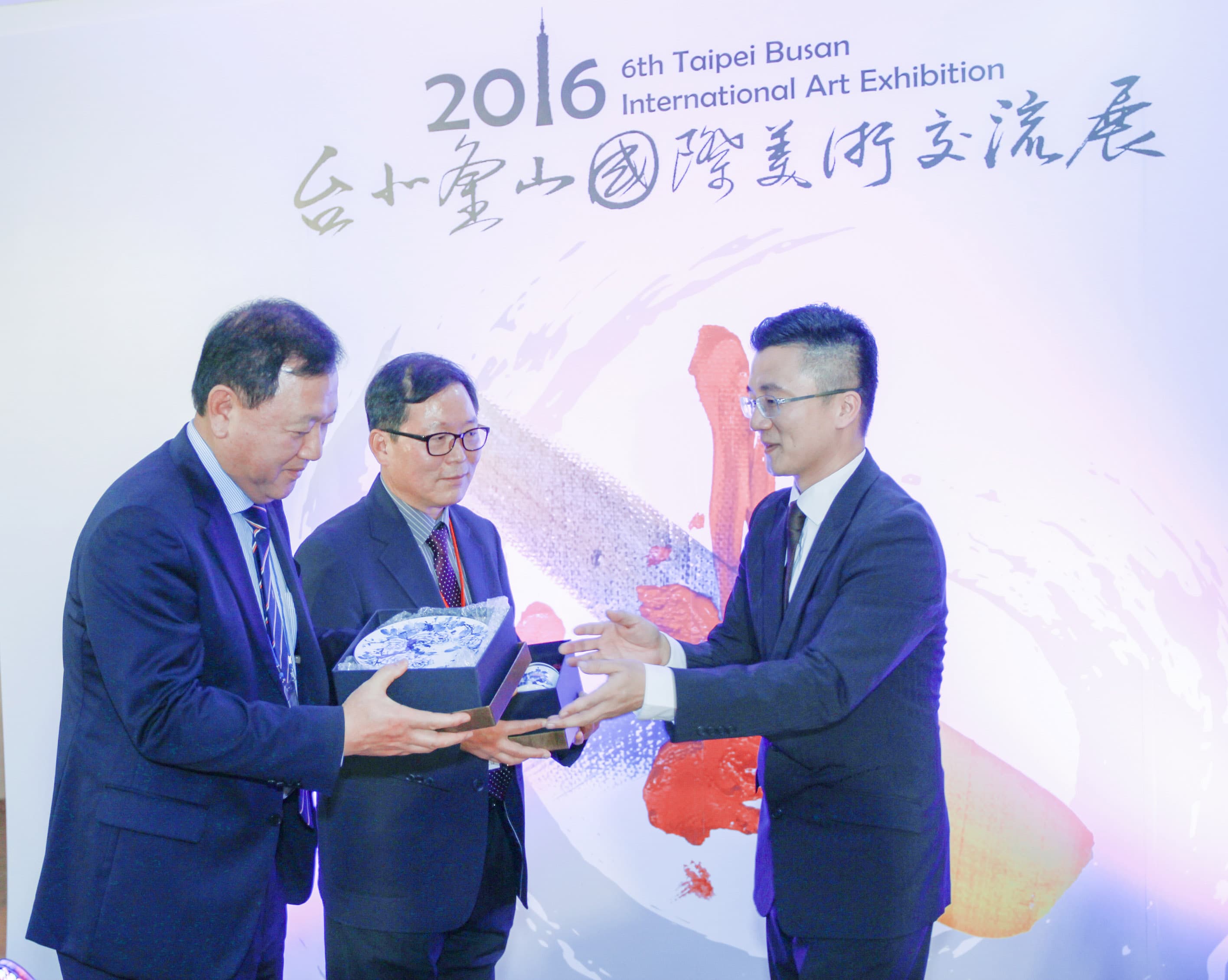 這張「2016年臺北釜山國際藝術展」的照片是三位男士站在展覽的背景前,其中兩位男士手持獎牌,照片的背景是展覽的標誌和裝飾。