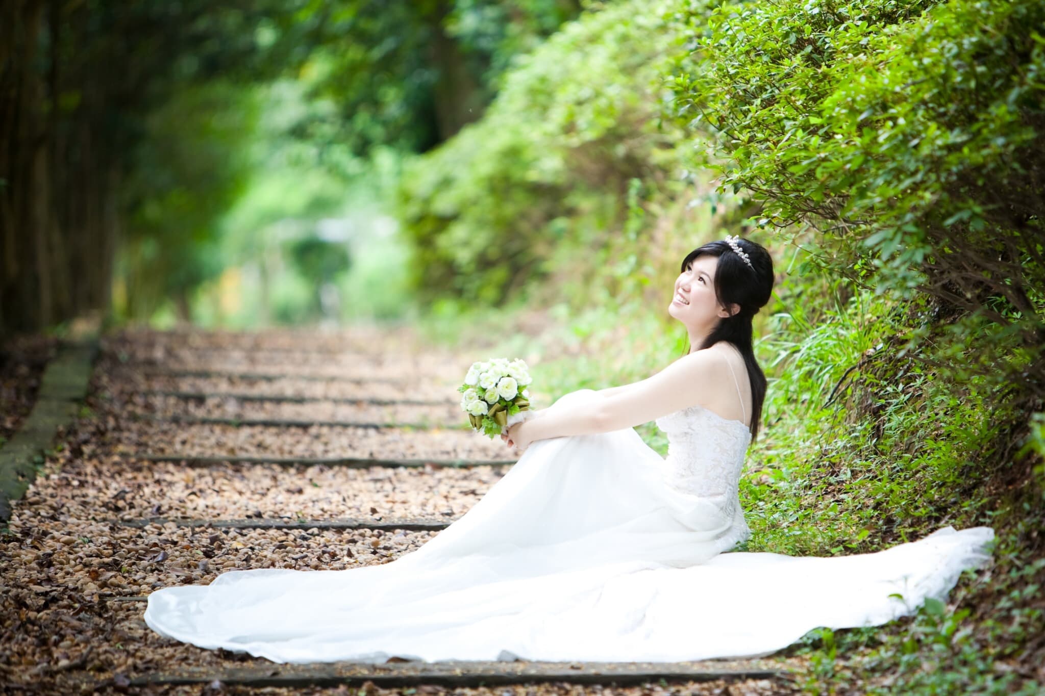 這張「婚紗照」的照片是一個穿著白色婚紗的女子,她坐在綠色草地上,笑容燦爛,手裡拿著一束白色的花,背景是一片綠色的樹林。