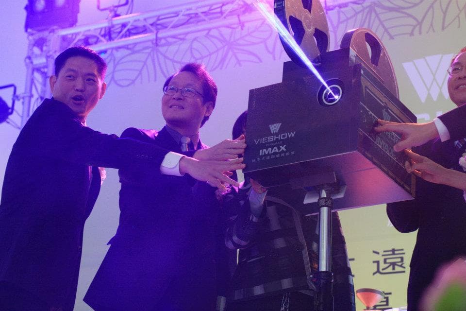 這張照片是三位男士在一個大型的 IMAX 影院投影機前合影,他們正對著鏡頭露出笑容,投影機上印有「VIESHOW IMAX」的字樣,是照片中最顯眼的元素。