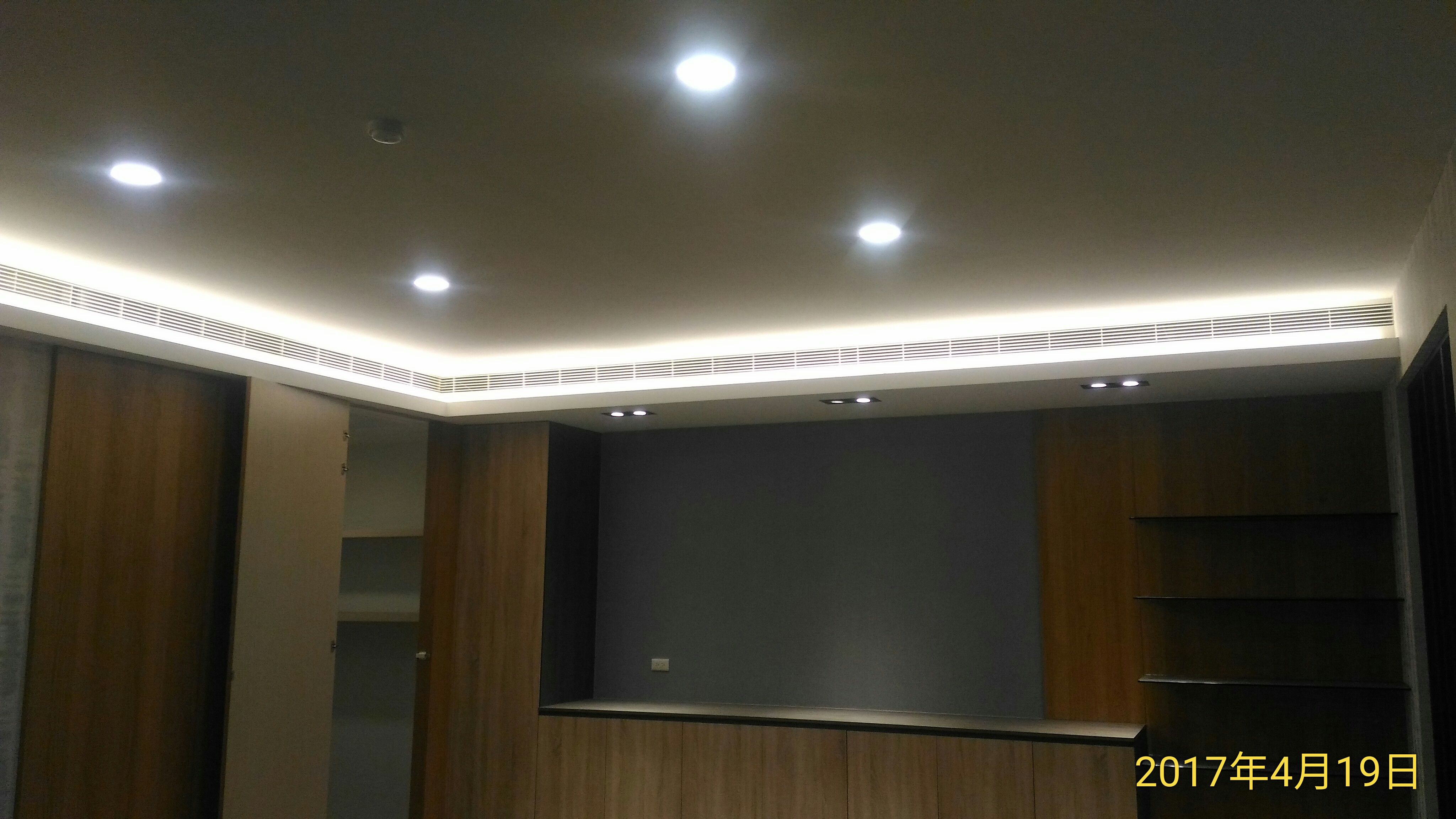 1) 照片顯示一個裝潢精緻的客廳,天花板上有嵌入式燈光,牆壁是淺灰色,地板是淺色木質地板,客廳中有一個木質電視櫃,櫃子上方裝有嵌入式燈光,客廳右側有一整排木質櫃子,櫃子上方有一個嵌入式燈光