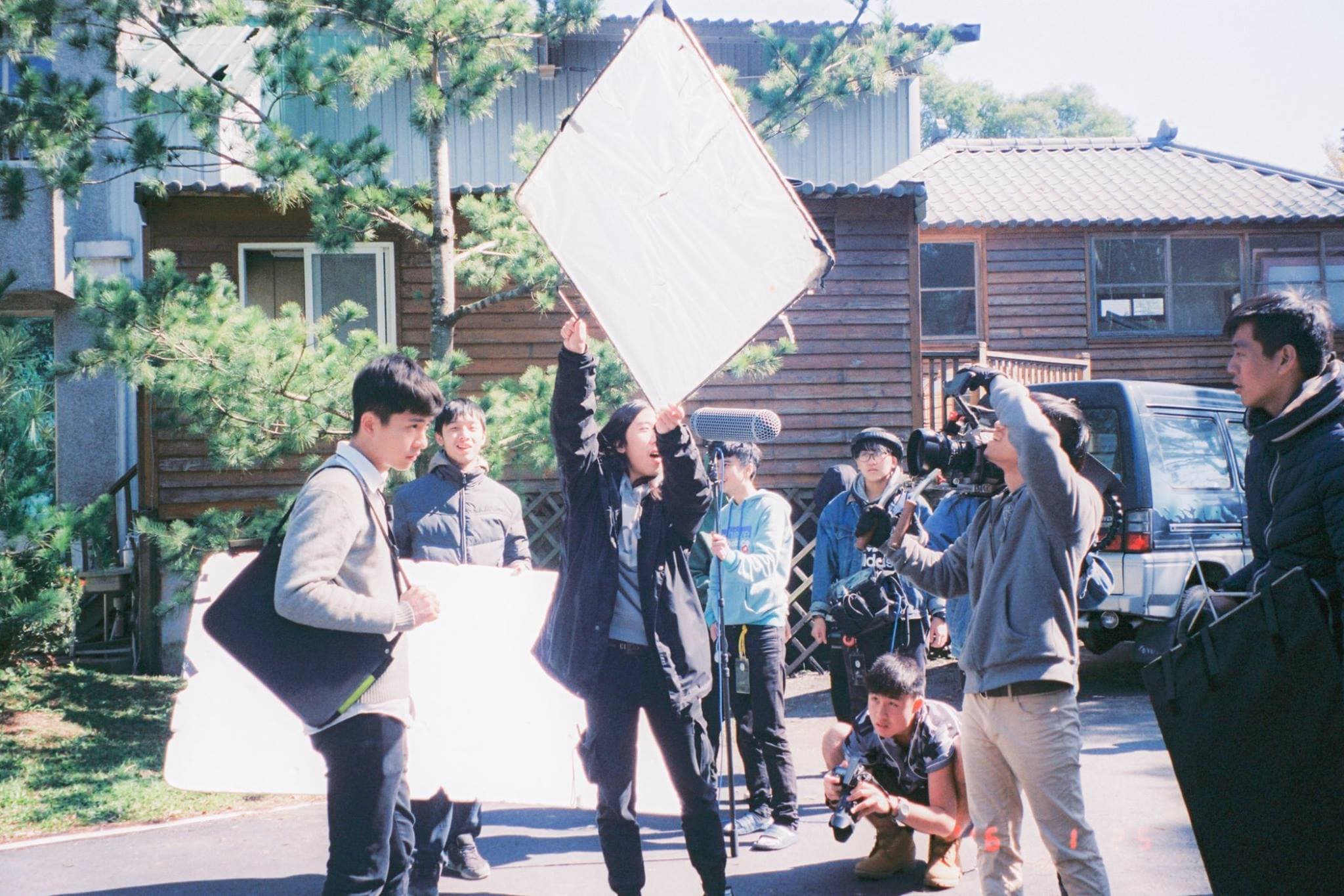 這張照片是一群人在戶外拍攝,其中最重要的是中間拿著相機的人,他正專注於拍攝,其他人則在旁邊協助或觀看。