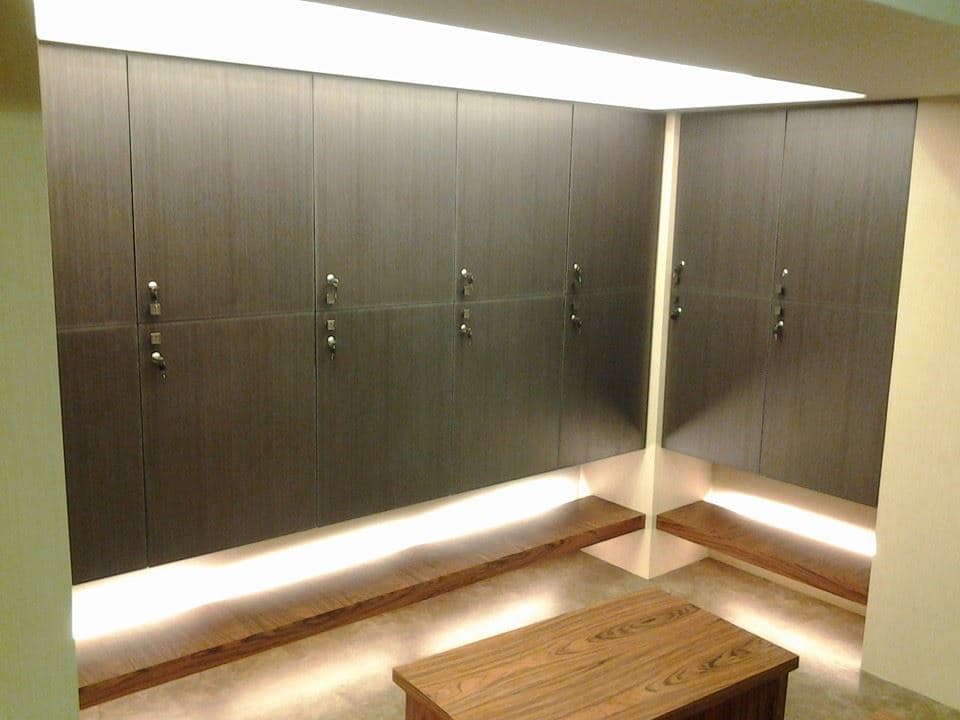 這張照片是一個現代化的更衣室,裡面有許多木質儲物櫃,櫃子上有燈光,牆壁上也有一些燈,整體感覺很明亮舒適。