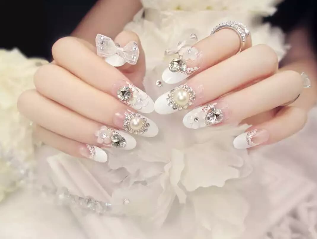 1) 照片顯示一雙戴著白色指甲油和裝飾著白色蝴蝶結、珍珠和鑽石的指甲的手