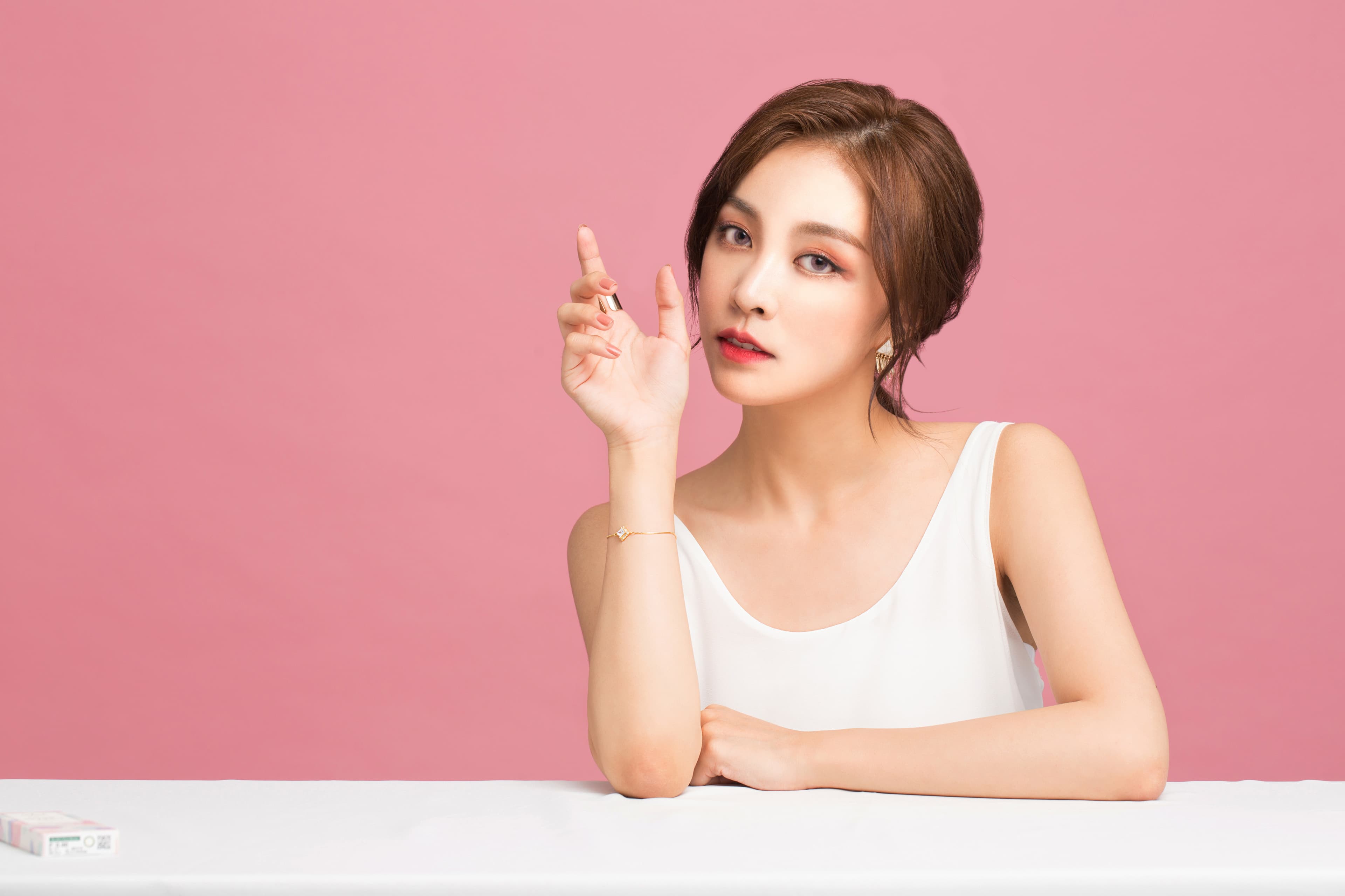 這張照片是一個年輕女子在粉紅色的背景下。她穿著白色背心,她的一隻手放在桌子上,另一隻手舉起來,指尖朝上。她看起來很自信,並且微笑地看著鏡頭。