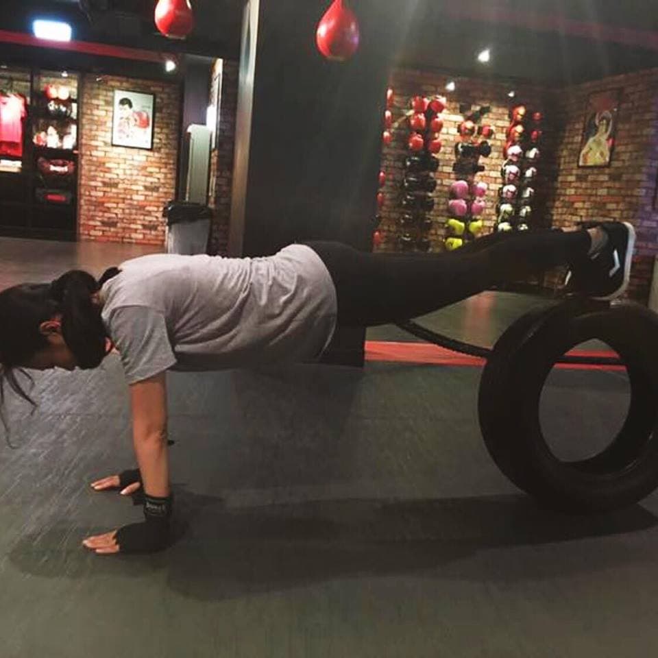 這張照片是一個女孩正在做平板支撐運動。她雙手撐在地板上,身體呈現一條直線,腿部抬高,腳放在一個黑色輪胎上。她專注於運動,表情認真。