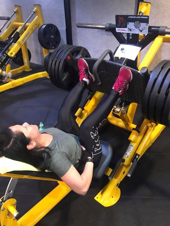 這張照片是一個女性在健身房使用腿部訓練器,她正努力鍛鍊腿部肌肉,她的表情專注而堅定。