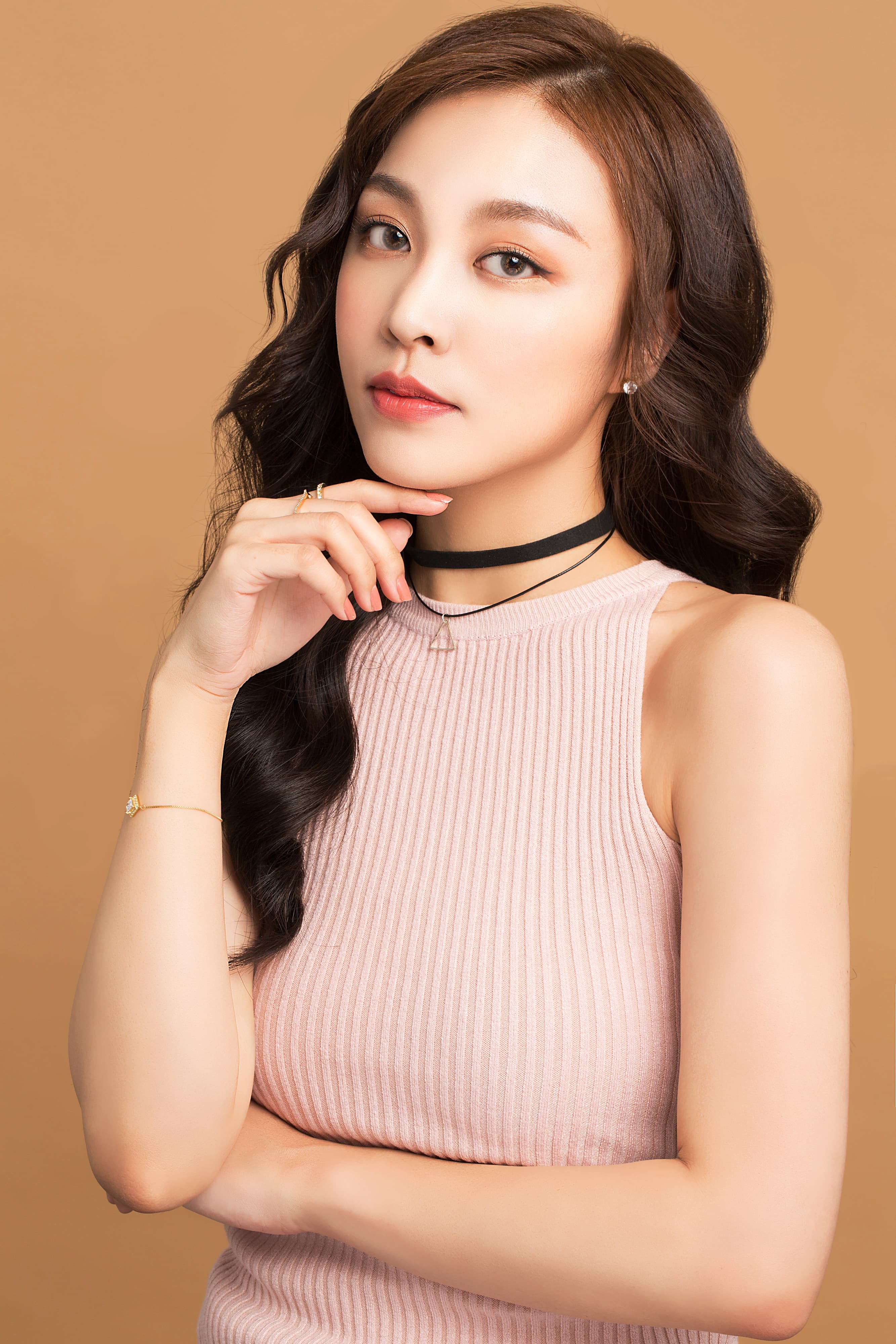 這張照片是一個年輕的女性,她穿著一件粉紅色的背心,手放在下巴上,眼神溫柔地看著鏡頭。她的五官精緻,頭髮烏黑亮麗,散發出優雅的氣質。