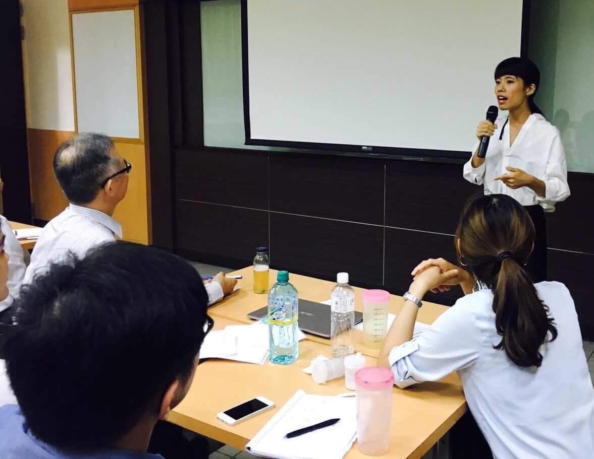 這張「會議」的照片是,一位女性正在臺上用麥克風演講,臺下有三位聽眾,其中一位坐在桌邊認真地聽著。