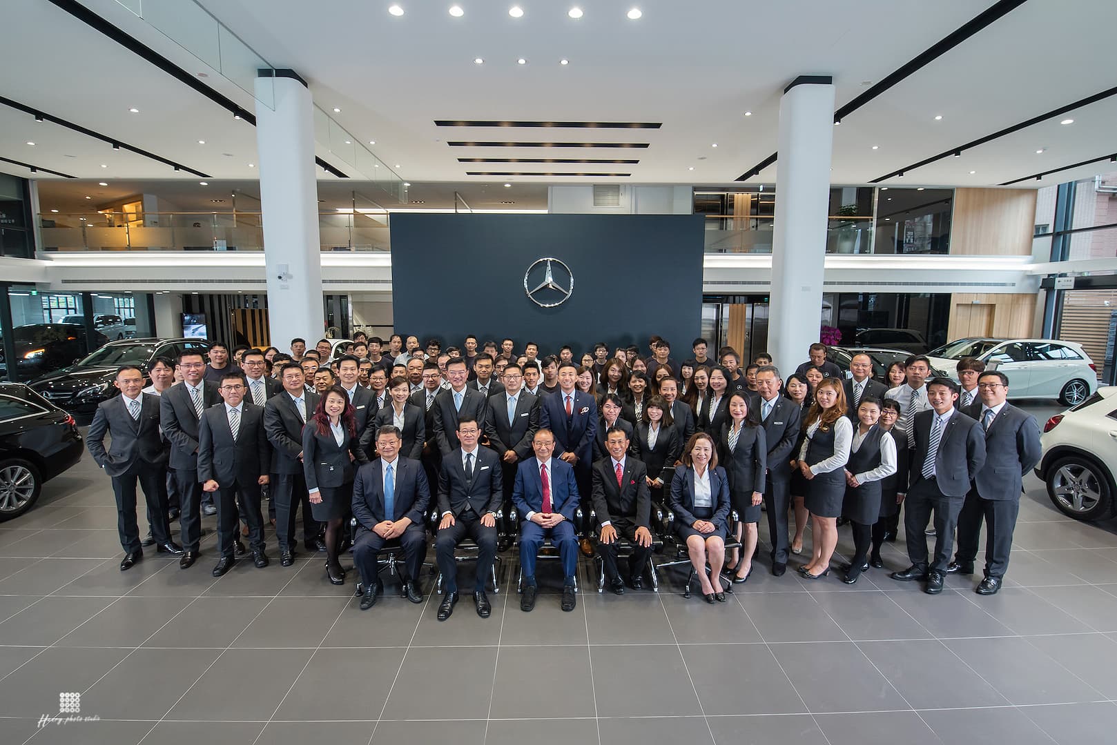 這張照片是一群人在汽車展廳裡合影。他們都穿著正式的服裝,並且笑容滿面,展現出友善和自信的氛圍。牆上有一個巨大的賓士標誌,這顯示這是一家賓士汽車經銷商,而他們可能是該公司員工或客戶。照片裡的每個人都面帶笑容,讓人感覺充滿了活力和希望。
